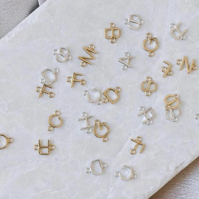 14k yellow gold and sterling silver initial connectors for permanent jewelry on a marble background. Letters are scattered, but include C, D, R, G, F, L, N, S, M and H.