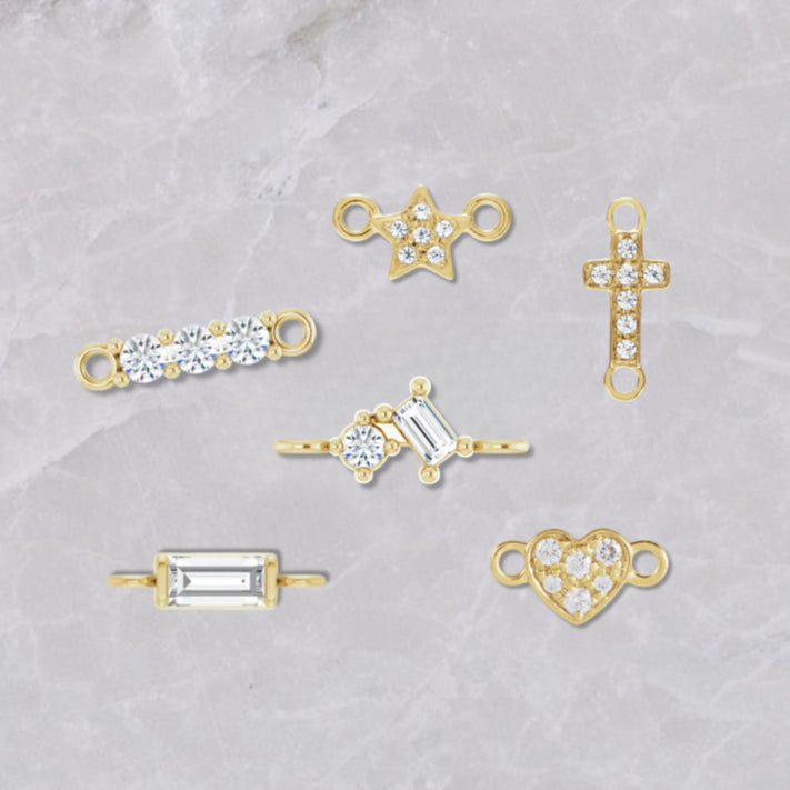 Six 14k gold permanent jewelry connectors on a white marble background. From left to right, the picture shows a triple diamond, baguette, double diamond, star diamond, cross diamond, and heart diamond connectors.