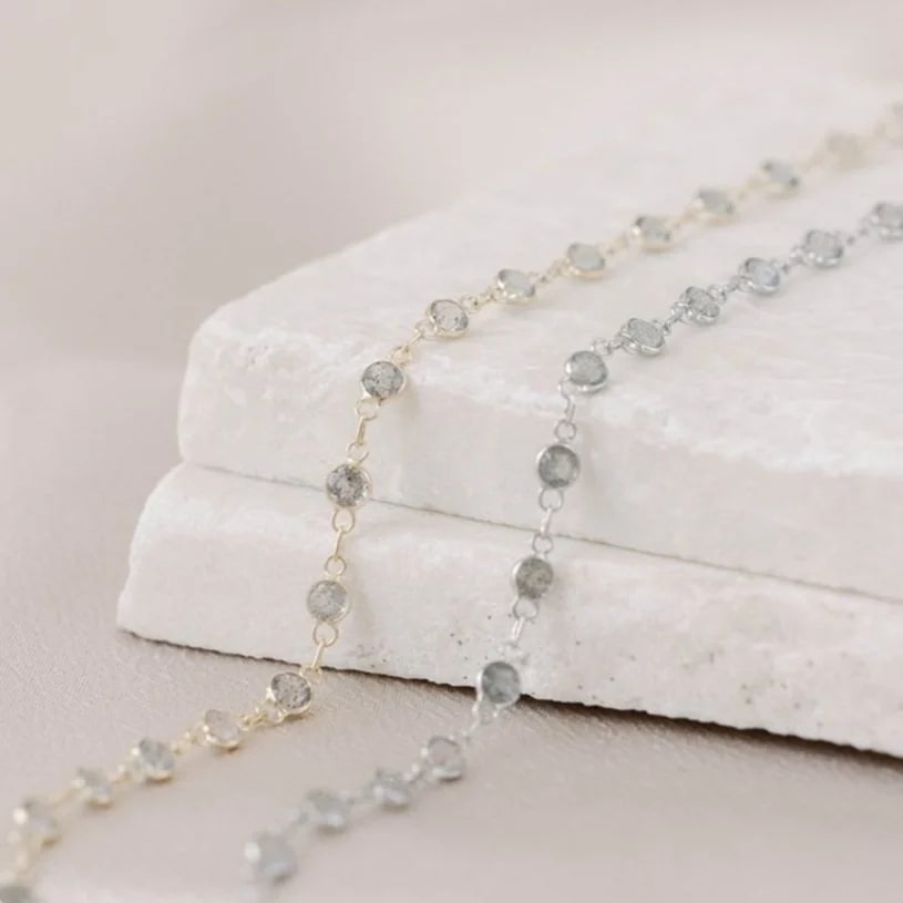 Yellow and white gold permanent jewelry chains, including three 14k circle links followed by a labradorite gem in a pattern. On a marble block and satin background. 