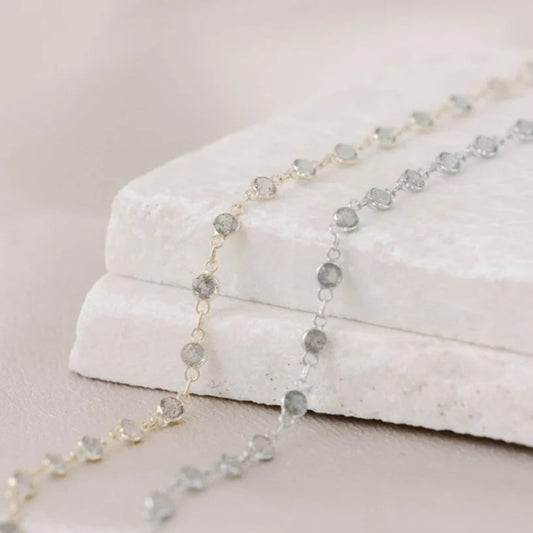 Yellow and white gold permanent jewelry chains, including three 14k circle links followed by a labradorite gem in a pattern. On a marble block and satin background. 