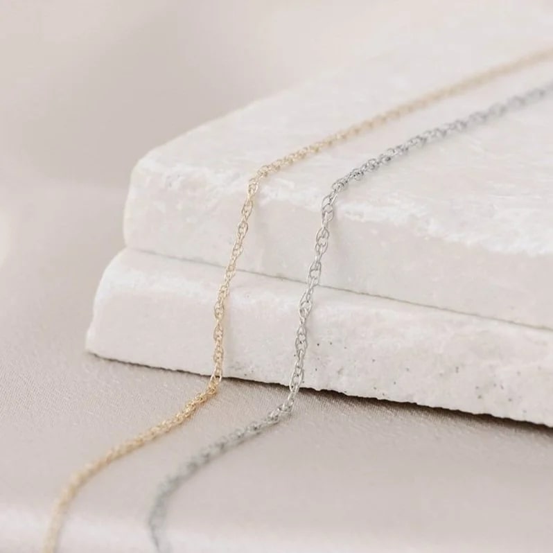 Dainty 14k white and yellow gold permanent jewelry chains laying on a stone surface with a neutral background.