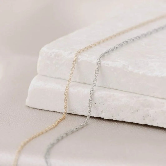 Dainty 14k white and yellow gold permanent jewelry chains laying on a stone surface with a neutral background.