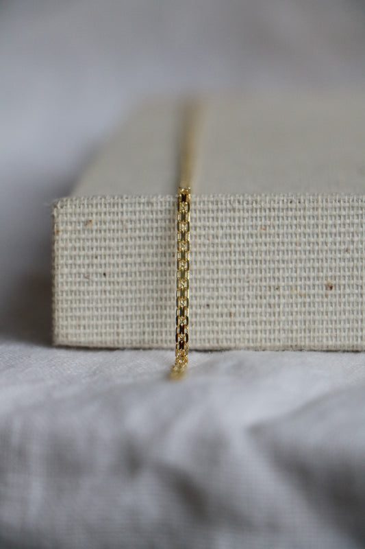 14k gold permanent jewelry chain, shown on a close up with square links and a flat 14k gold piece across the middle of each link. Sitting on a woven jewelry holder. 
