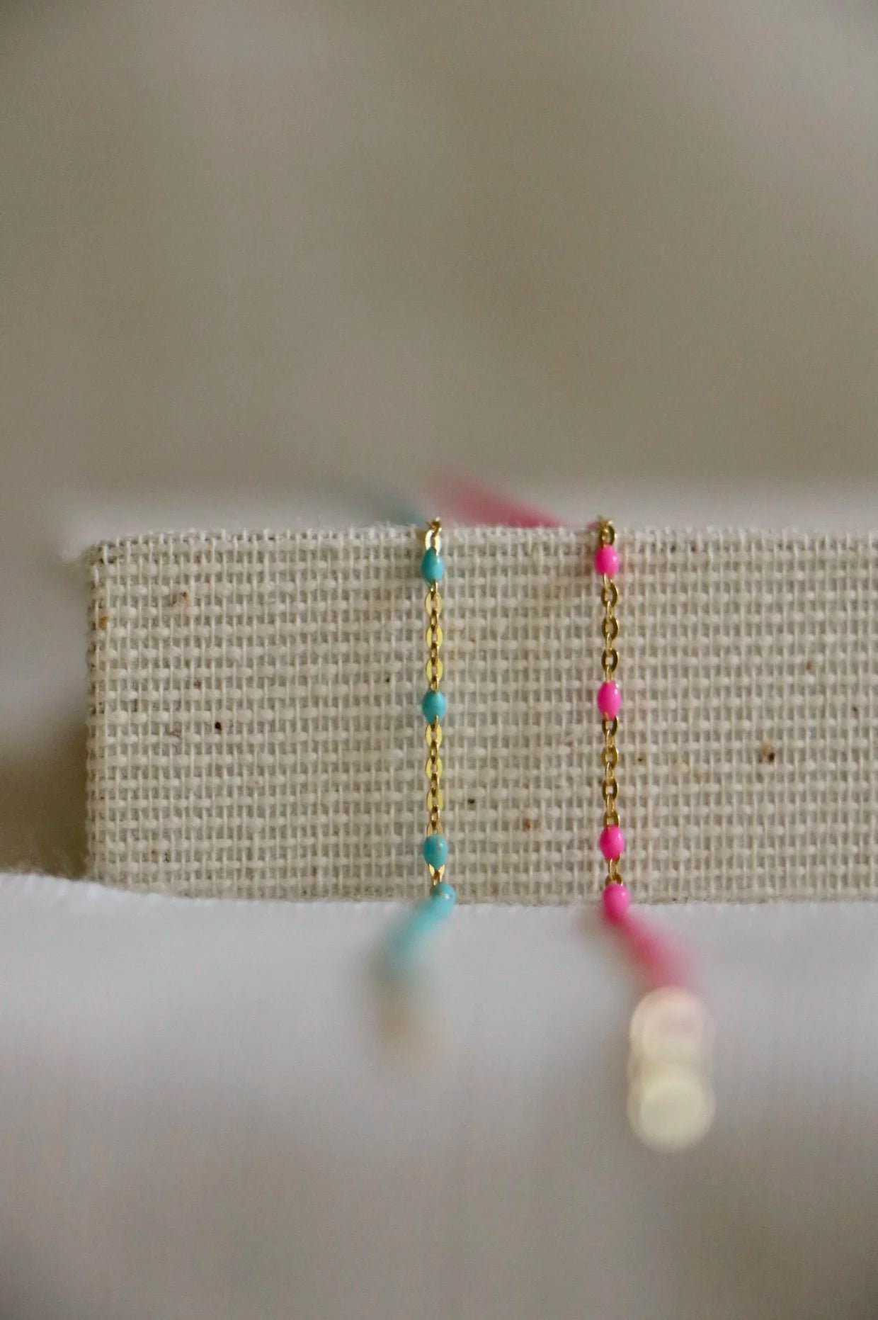 Two 14k gold permanent jewelry chains on a woven holder. The left is a turquoise option, the right a hot pink option. The chains have three gold links followed by a color pop link in a pattern.