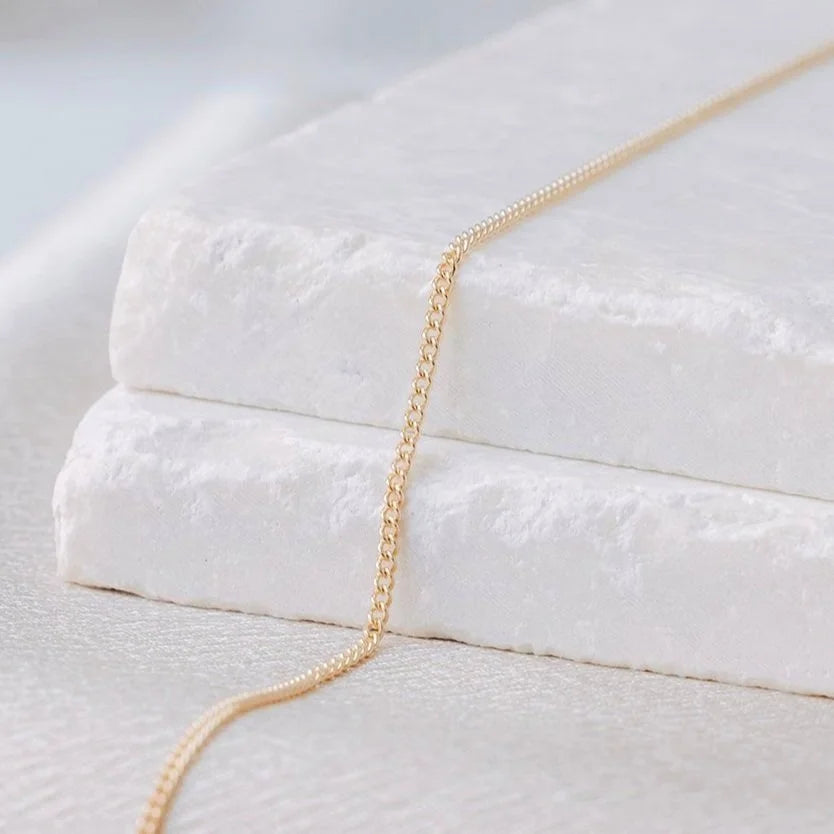 Dainty 14k gold Curb Link from Charm City Links laying on a white stone surface with a blurred background.