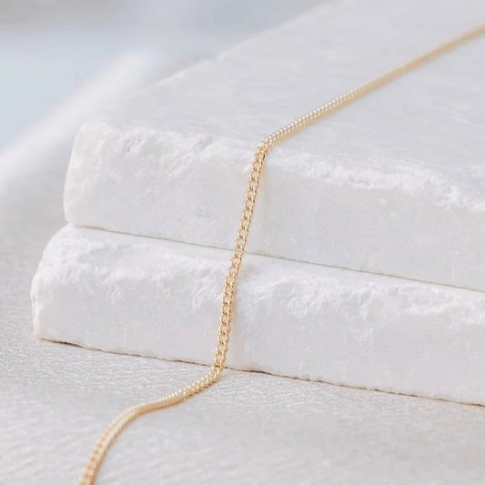 Dainty 14k gold Curb Link from Charm City Links laying on a white stone surface with a blurred background.