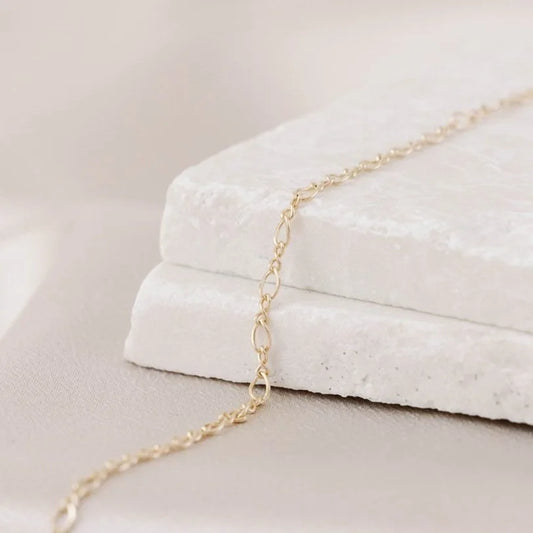 14k gold permanent jewelry chain in the shape of a figure eight or infinity symbol, laying on a neutral blurred background.