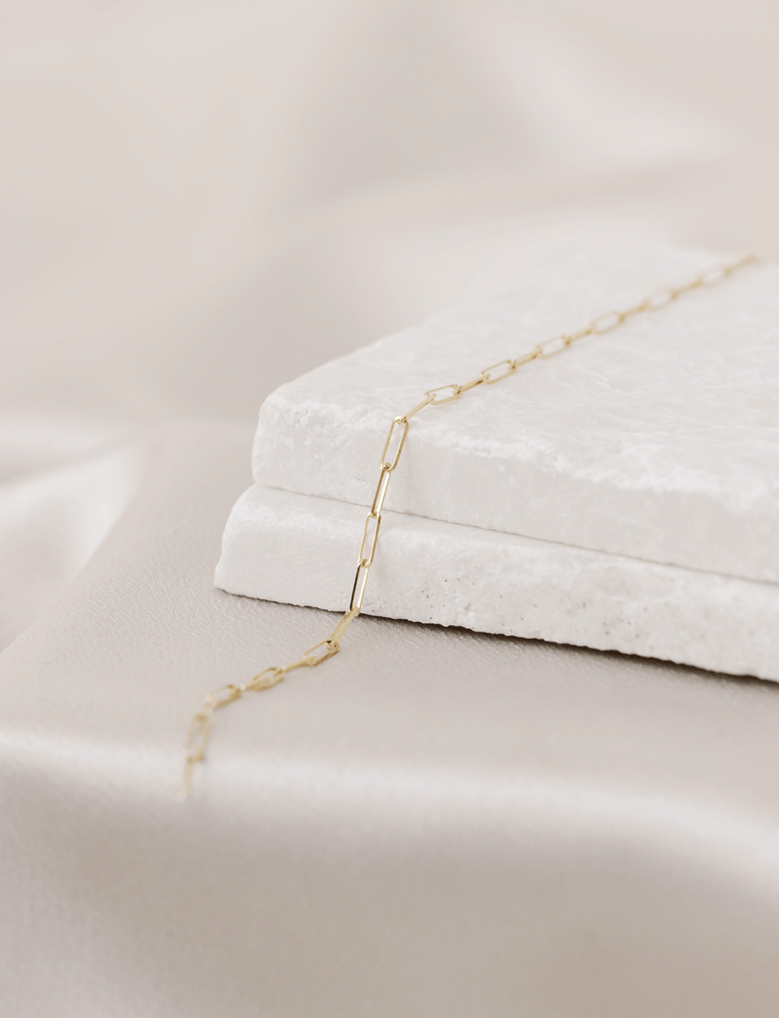 Classic 14k gold permanent jewelry chain with a paperclip design, dainty, and laying on two stones with a silk background. 