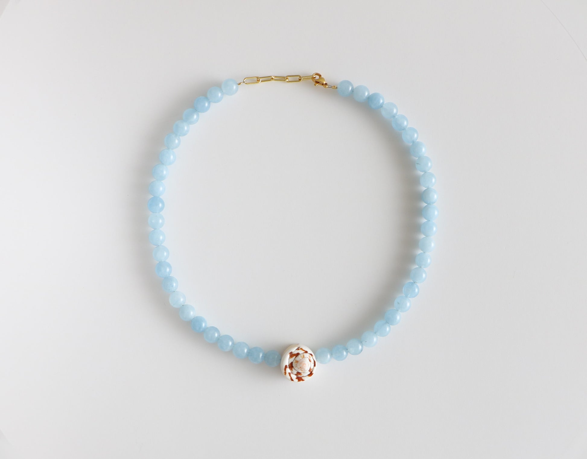 Beaded shell necklace, measuring 18'' in length with light blue beads around, a brown and white swirled shell at the bottom, and a gold clasp. Laying flat on a white background.