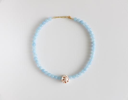 Beaded shell necklace, measuring 18'' in length with light blue beads around, a brown and white swirled shell at the bottom, and a gold clasp. Laying flat on a white background.