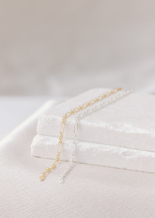 Two permanent bracelet chains on a light stone and white background. This shows Charm City Links Oval Link, with small circular connectors and large oval links alternating in a pattern. Shown on left in gold-filled, on right in sterling silver.