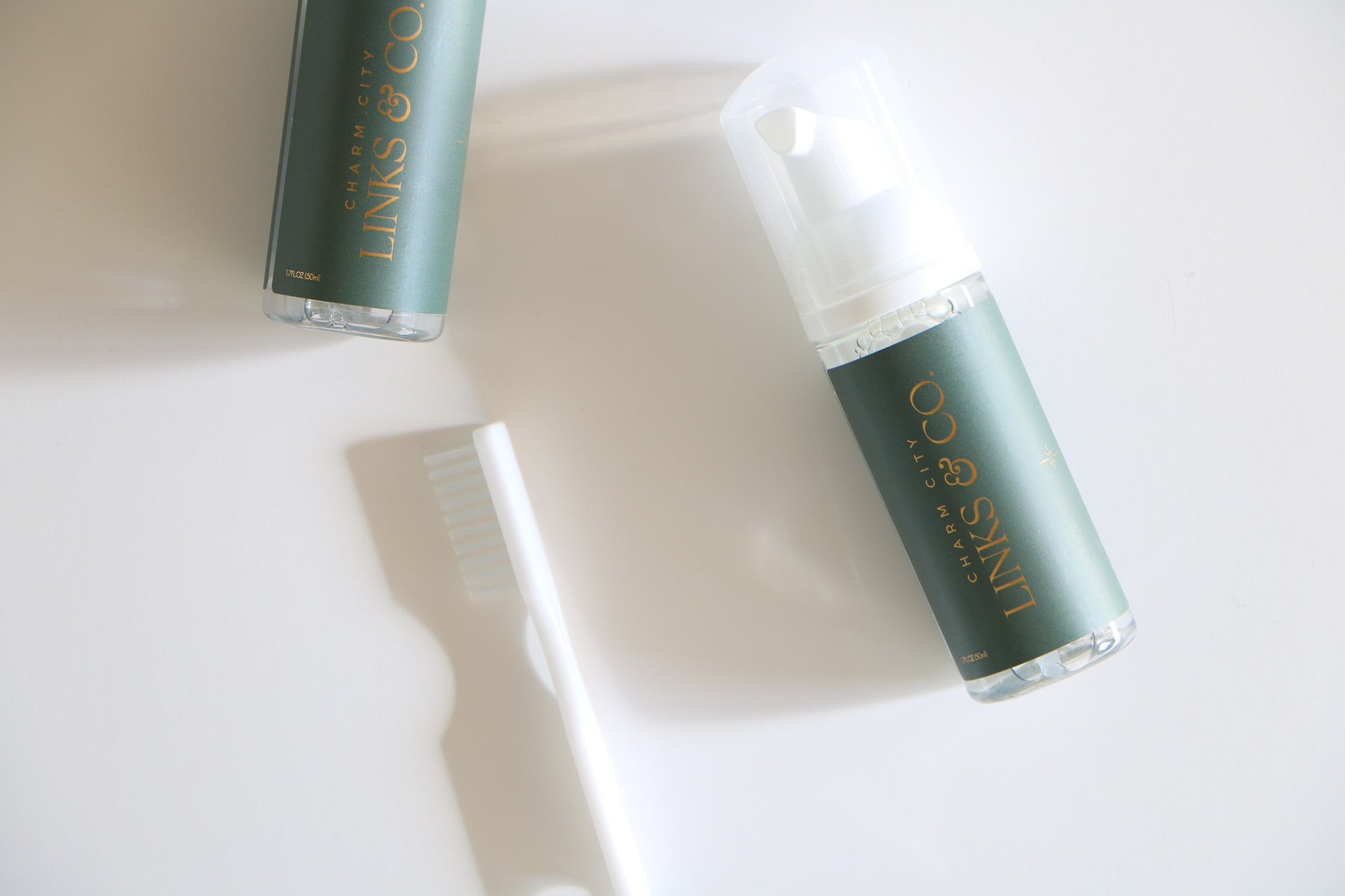 Overhead view of Charm City Links permanent jewelry cleaner for their sterling silver, gold-filled, and 14k gold jewelry. Two bottles have a dark green label with their gold logo, and includes a mini scrub brush.