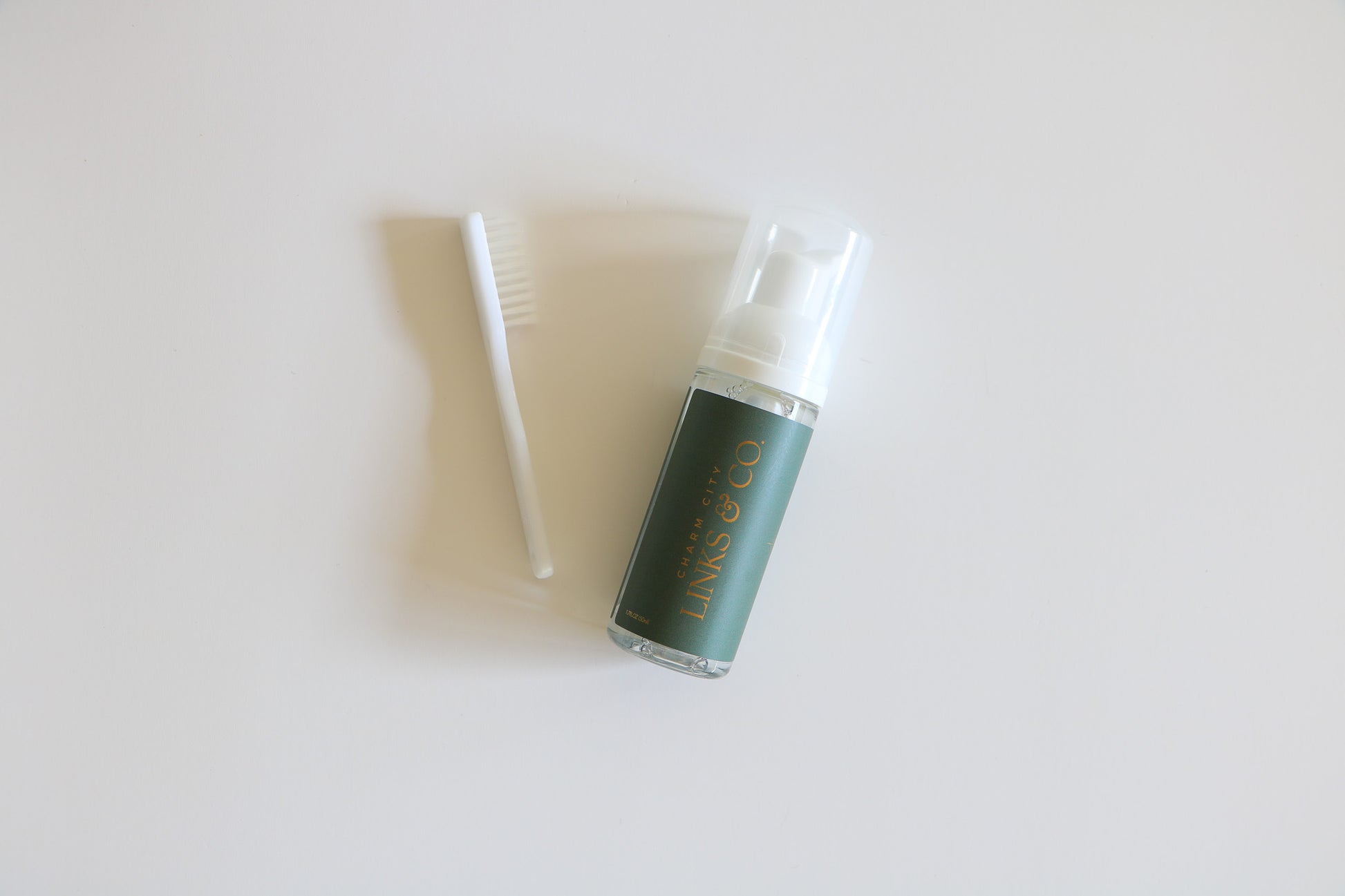 Overhead image of single Charm City Links permanent jewelry cleaner bottle. The dark green label wrap with their gold logo has a pump top, and a white mini scrub brush is to the left, included with purchase.