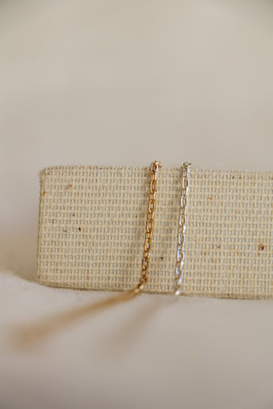 Two cable link permanent jewelry chains, the left in gold-filled and the right in sterling silver, sitting on a woven holder with a blurred white background. 