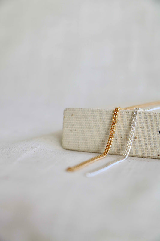 Two permanent jewelry Cuban links, one gold-filled and the other sterling silver, laying across a woven surface with a blurred white background.
