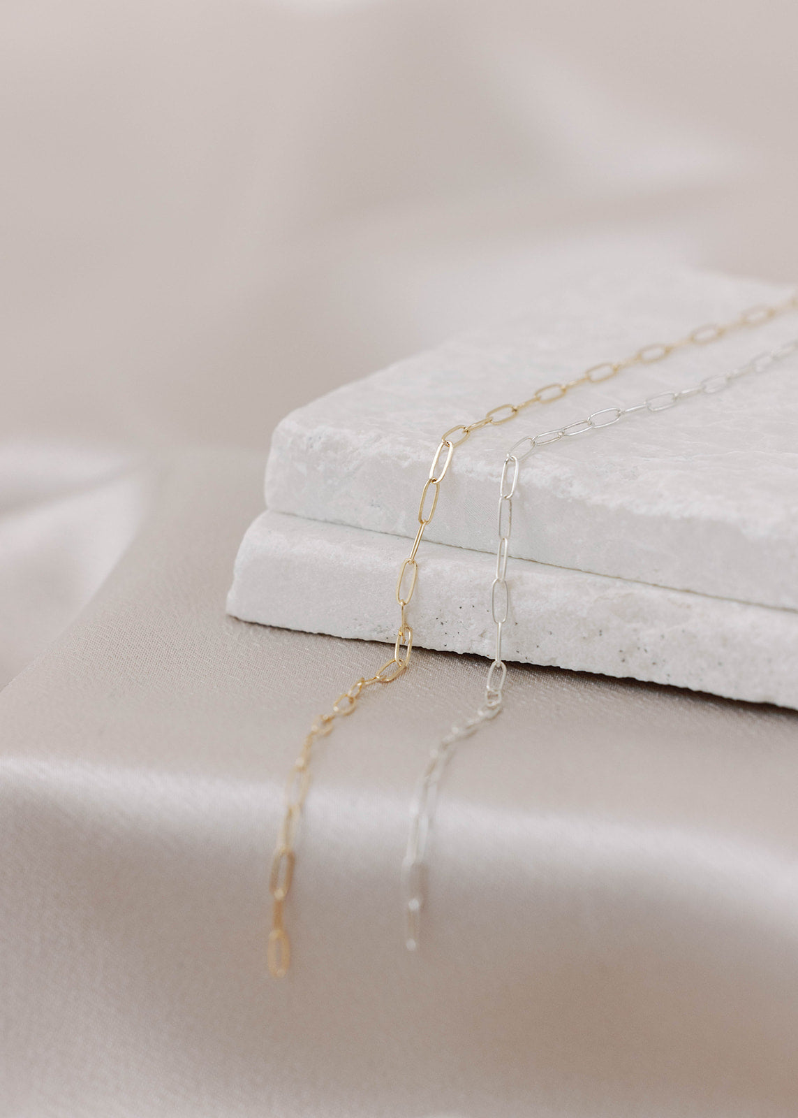 Dainty paperclip permanent jewelry links on a stone sitting on top of a champagne satin tablecloth. Gold-filled and sterling silver shown. 