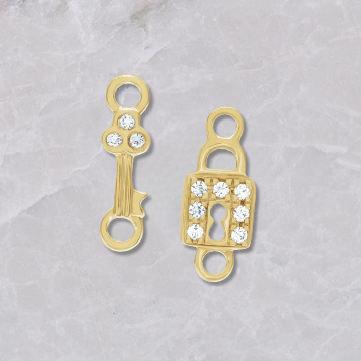 Two 14k yellow gold and diamond permanent jewelry connectors on a marble background. The left has a gold key with three diamonds on top, the right has a lock with 7 diamonds around the square lock. 