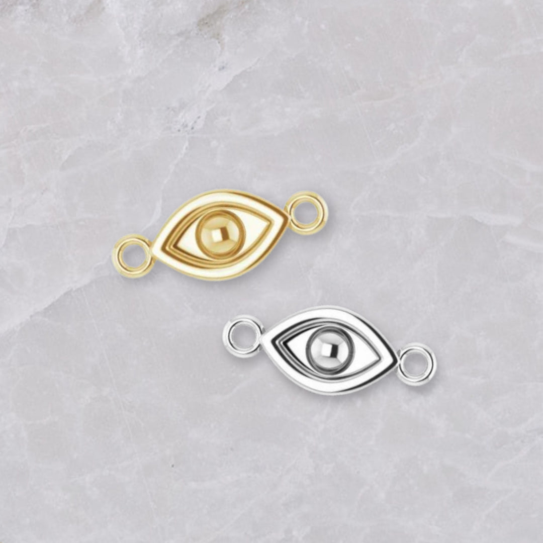 Two permanent jewelry connectors on a marble background. In the shape of an evil eye with connectors on the left and right, they are show in 14k yellow gold and sterling silver.