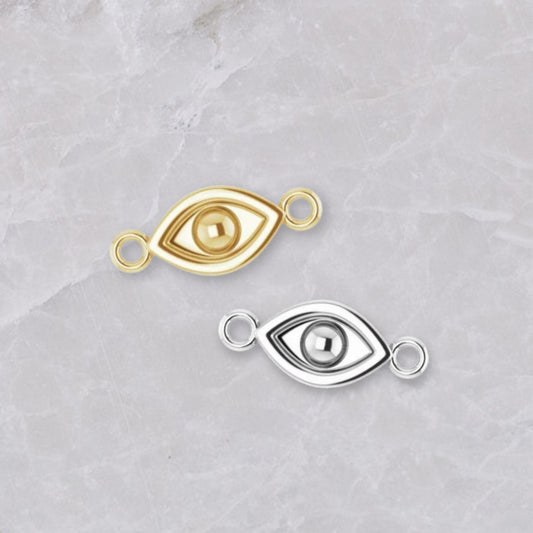 Two permanent jewelry connectors on a marble background. In the shape of an evil eye with connectors on the left and right, they are show in 14k yellow gold and sterling silver.
