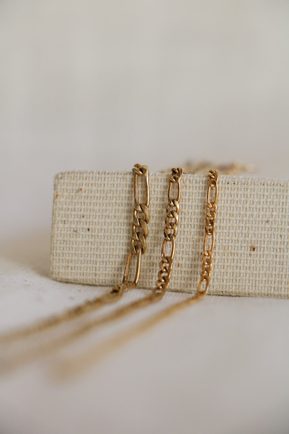 2mm, 3mm, and 4mm gold-filled Figaro Link permanent jewelry chain, draped side by side on a woven jewelry holder. They go from largest to smallest, including three small circular links followed by a large oval link in a pattern.
