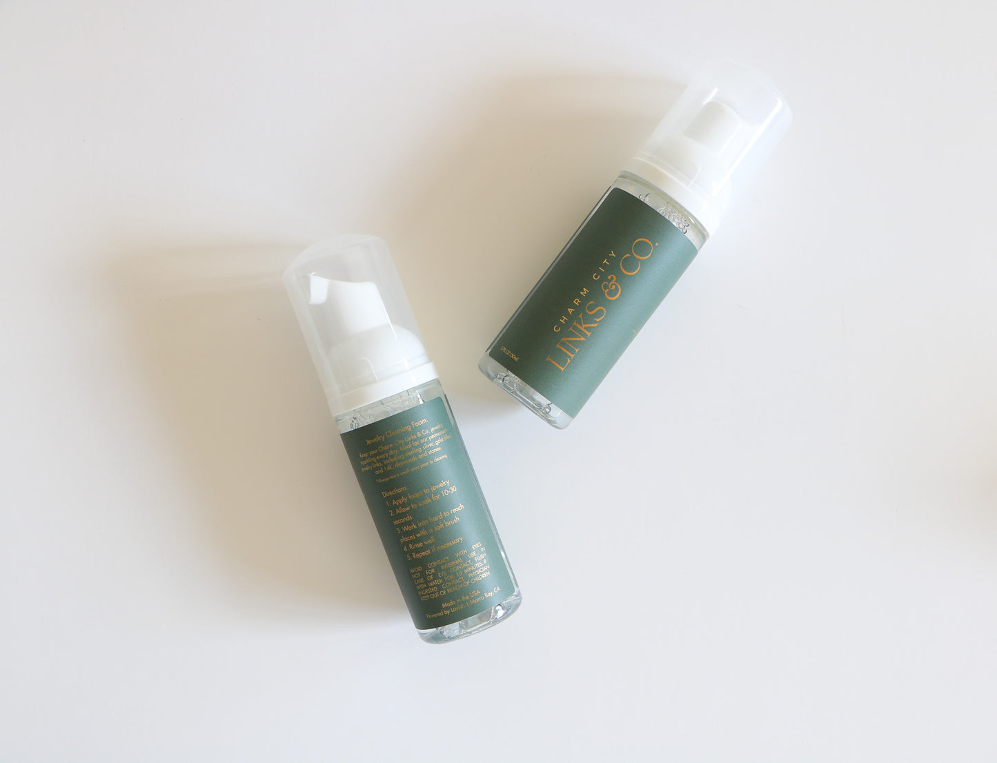 Charm City Links jewelry cleaner bottles. Two bottles shown diagonally, with one showing the back instructions and the other the front logo. Forest green label with gold logos. 