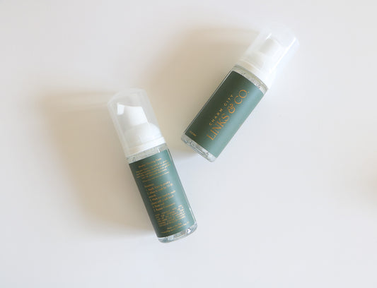 Charm City Links jewelry cleaner bottles. Two bottles shown diagonally, with one showing the back instructions and the other the front logo. Forest green label with gold logos. 