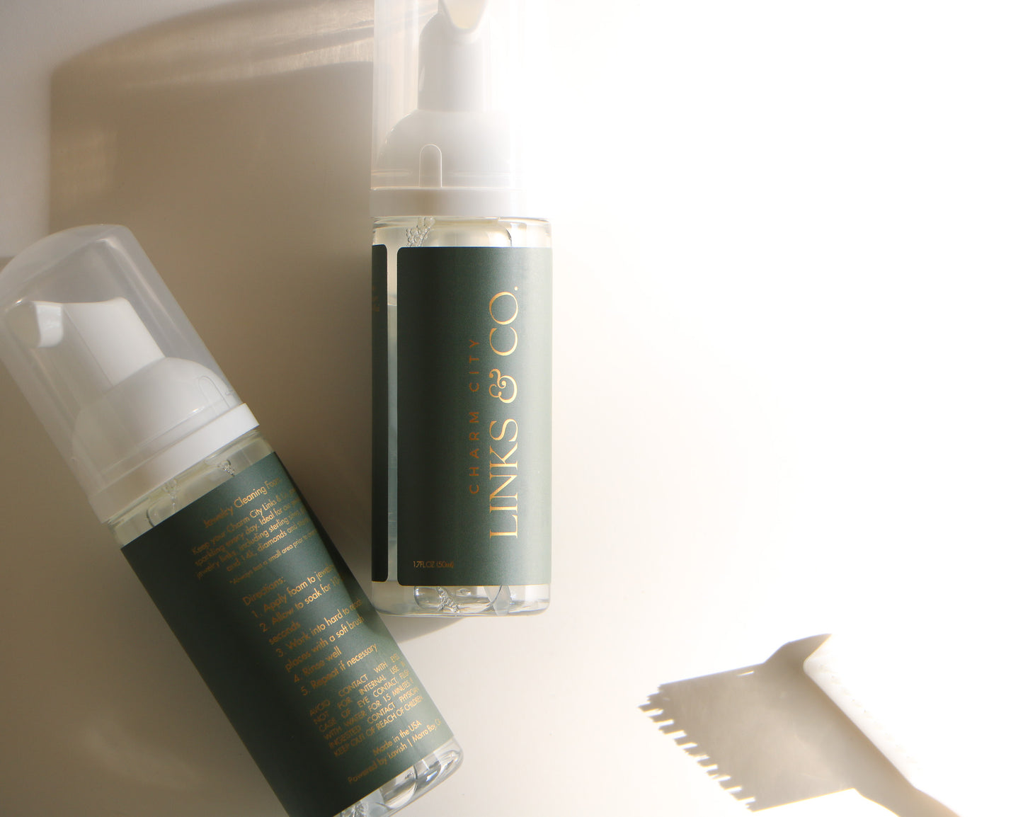 Sun and shadow close-up of Charm City Links permanent jewelry cleaner. It shows two bottles, the left with the back label, the right with the front. The label wrap itself is a deep green, with gold font and logo. 