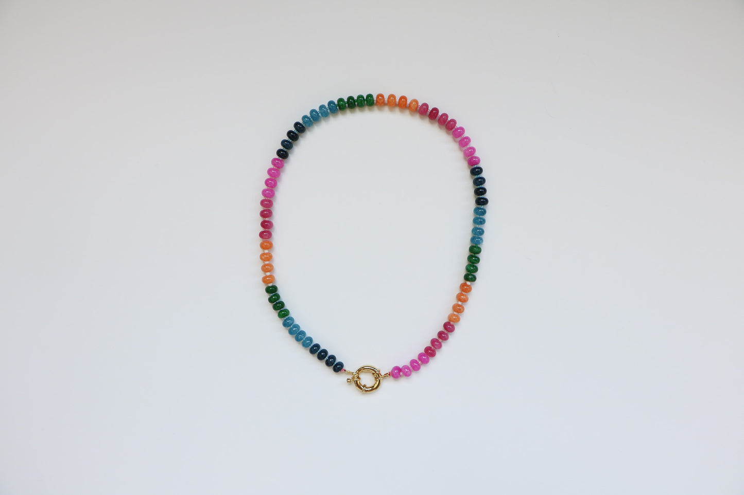 Natural gemstone beaded necklace in the color 'Candy'. Rainbow patterned beads, including dark blue, light blue, green, orange, red, and pink, connected in a 20'' necklace by a thick gold clasp, sitting on a white background. 
