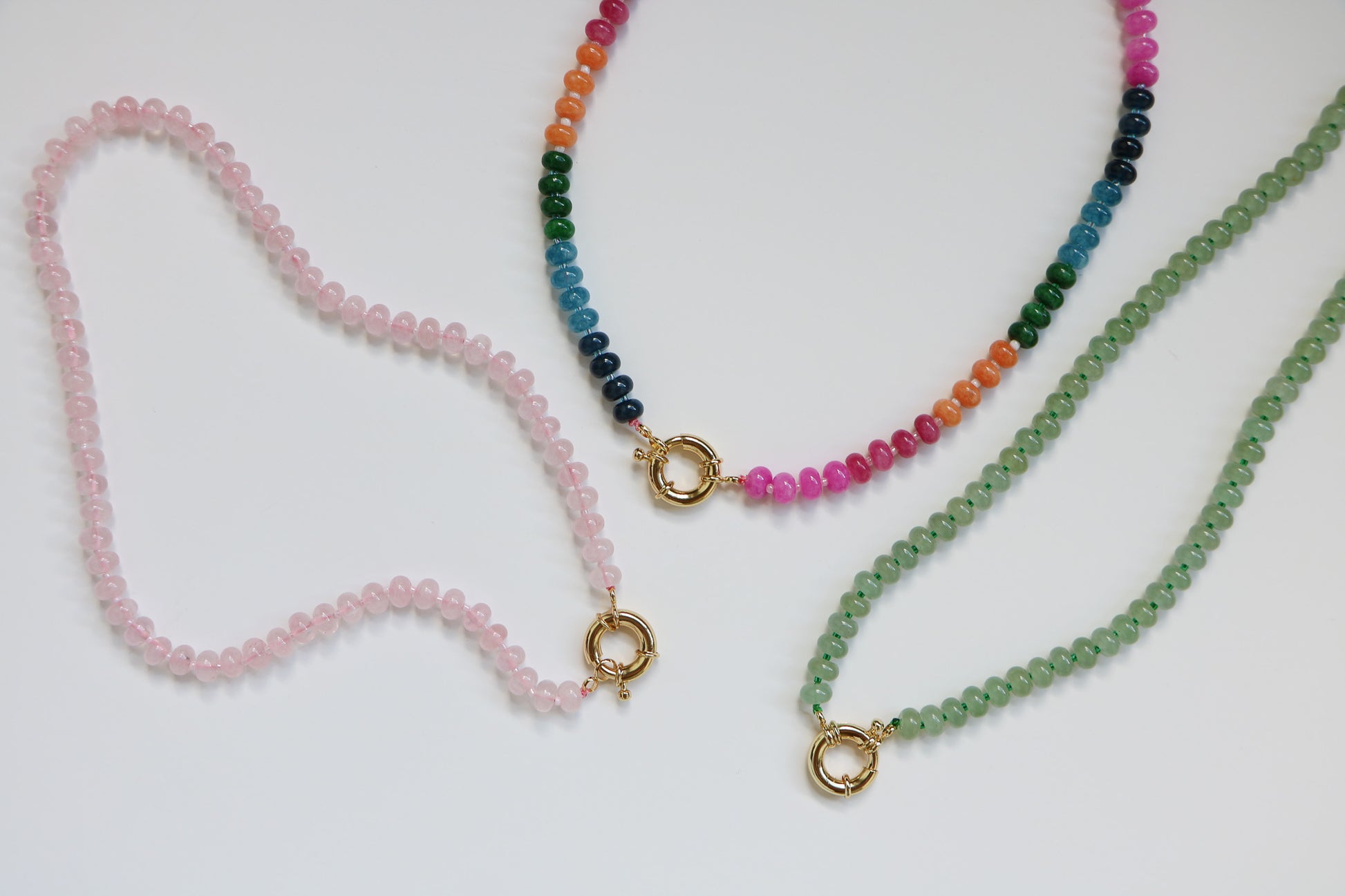Flatlay of three natural gemstone necklaces from Charm City Links, called 'The Harper Necklace'. They are each about 20'', finished with a thick gold clasp. The left is in the color Bubblegum (light pink), the middle Candy (rainbow), and the right a light green. 