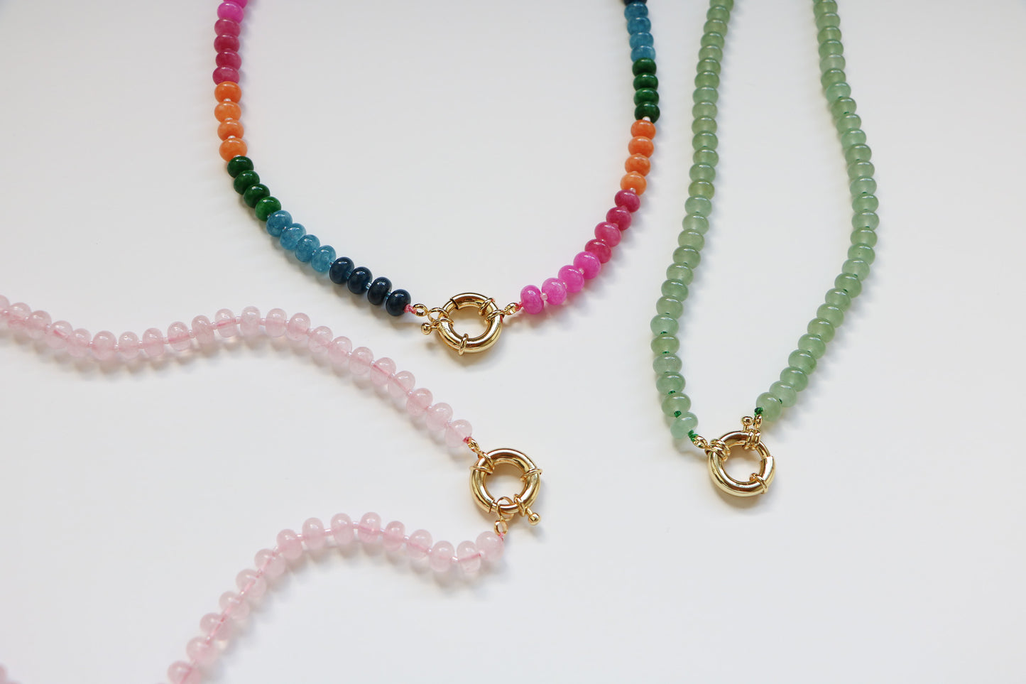 Three natural gemstone beaded necklaces  laying on a white background. All are connected by a thick gold clasp. The left is a bubblegum color, the middle is their 'candy necklace' option of rainbow beads, and the right is a seafoam green color. 