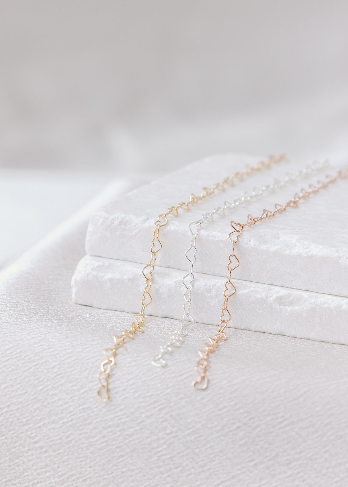Permanent jewelry links on a stone background and white satin sheet. The Heart Link has hearts in a line pattern, in gold-filled, sterling silver, and rose gold. 