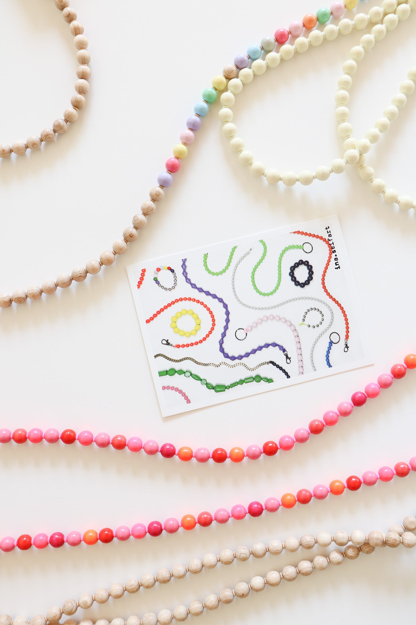 Close ups of Ina Seifart beaded lanyards, with parts of the pastel and cream ones at the top, and multicolored pink and neutral at the bottom, with a branded card in the middle of the flatlay. Made with wooden threaded beads.
