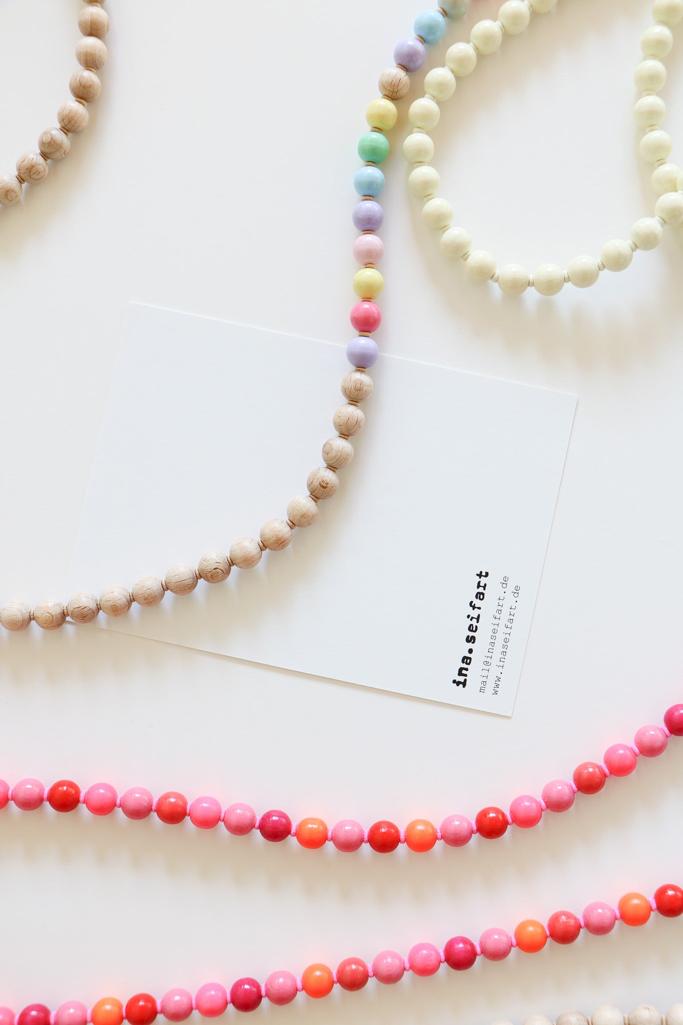 Close up of two threaded wooden beaded lanyards, one with neutral and pastel beads, the other with pink, red, and orange multicolored beads. An Ina Seifart branded card is sitting in the middle of the flatlay. 