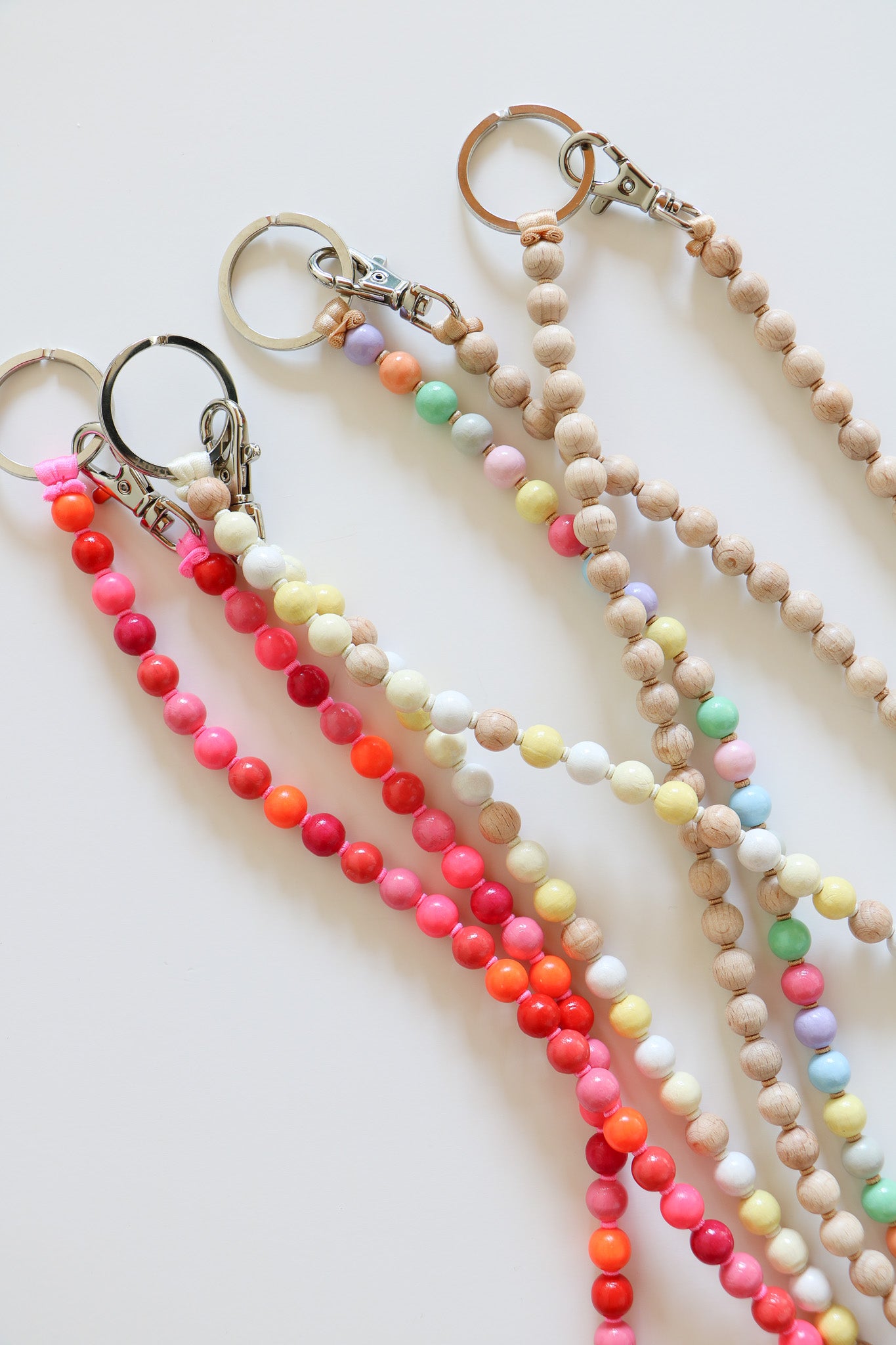 Colorful beaded keychains laying on a white background. These show wooden beaded lanyards with metal clasps, with options including pink, pastel, neutral, and cream.