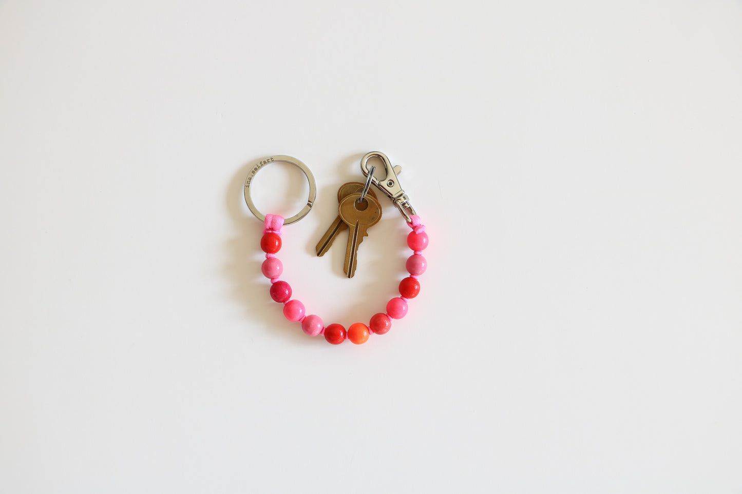 Pink multi-colored natural wood keychain arranged in a semi-circle. Metal ring on one side, metal clasp and two small brass keys on the other. Beads are light and medium pink, red, and orange. 