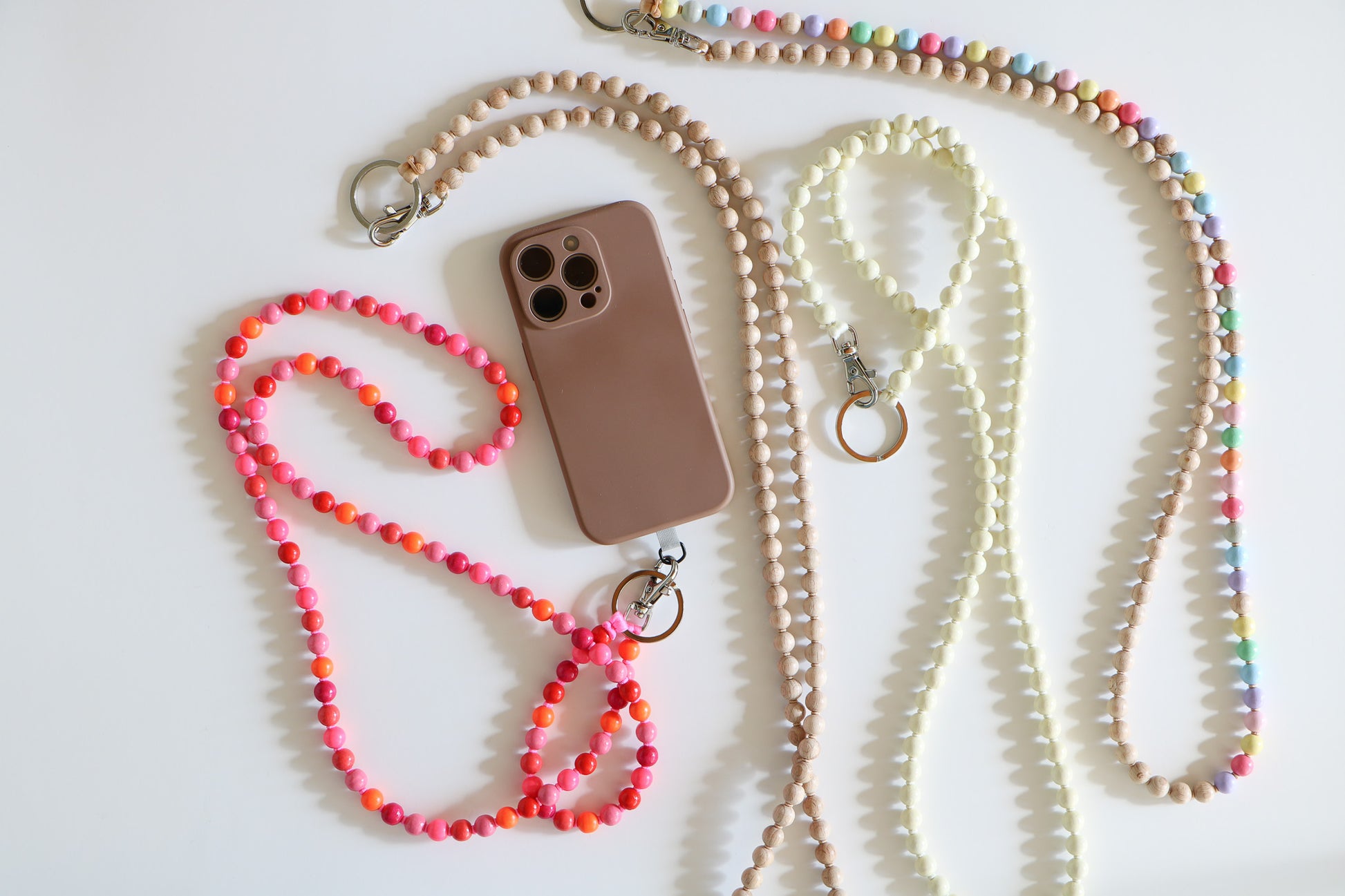 Multiple beaded lanyards in a flatlay. Wood threaded beads with a metal clasp, from left to right there is a multicolored pink lanyard, a neutral lanyard, a cream lanyard, and a rainbow lanyard.