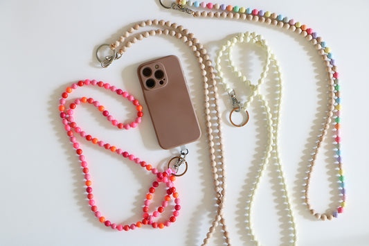Multiple beaded lanyards in a flatlay. Wood threaded beads with a metal clasp, from left to right there is a multicolored pink lanyard, a neutral lanyard, a cream lanyard, and a rainbow lanyard.