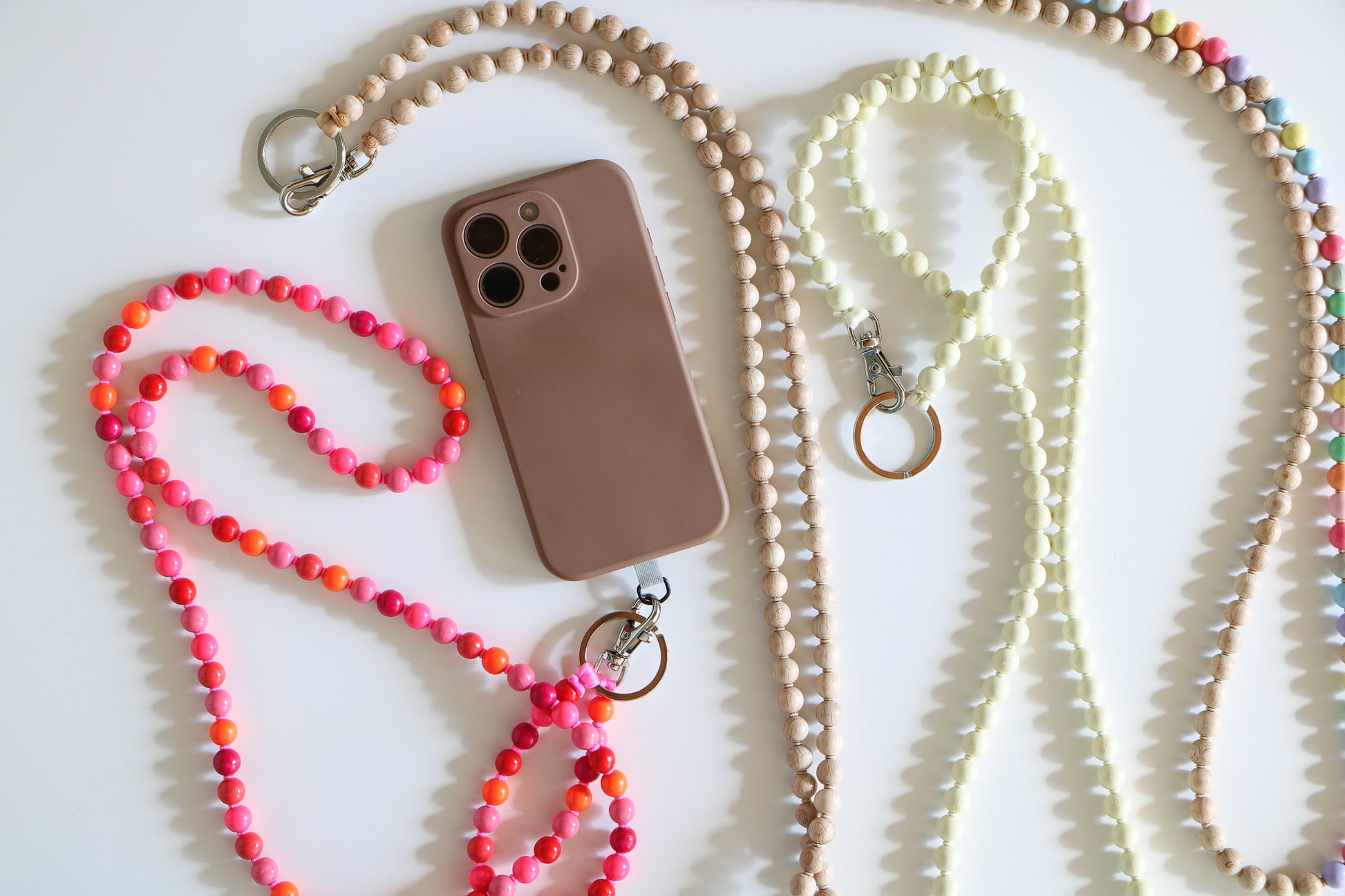 Flatlay of wooden beaded lanyards for phone or keychain, connected with a metal clasp. Offered in Pink Mix, Pastel Mix, Opal White, and Natural.