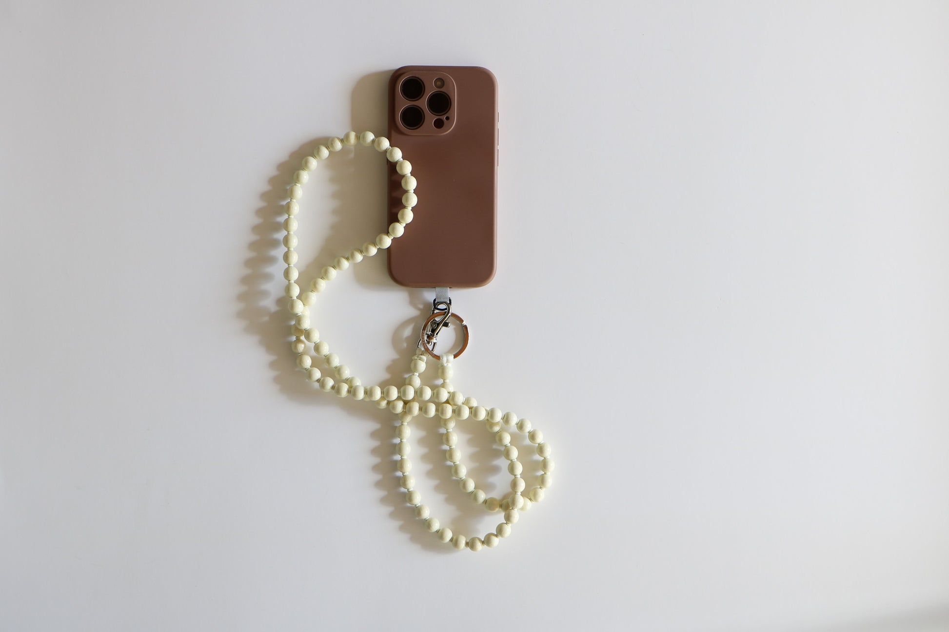 Phone with brown case face down on a white background, with a wooden beaded phone lanyard attached. The lanyard beads are an opal white color. 