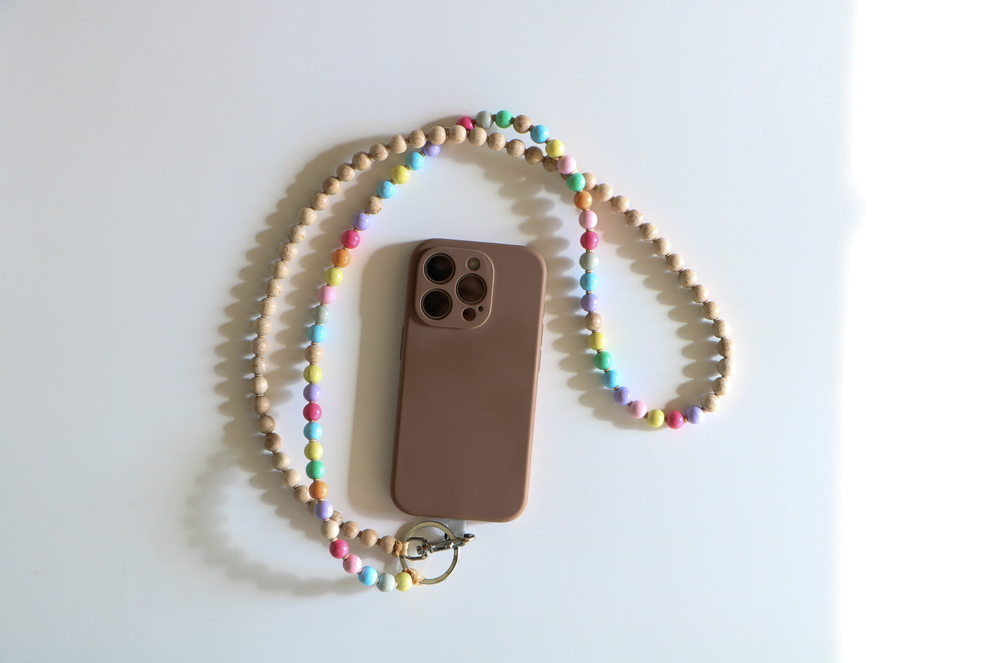Wooden beaded lanyard connected to phone with a metal clasp. Made by Ina Seifart, the lanyard is a mix of pastel and natural wood beads on a white flatlay.