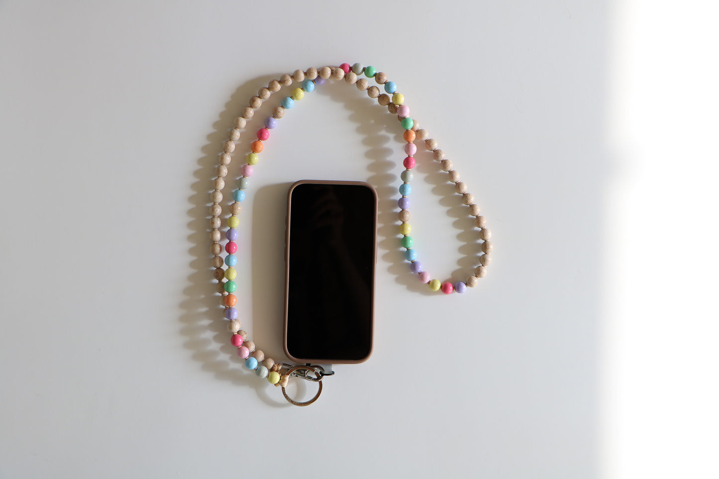 Phone with colorful beaded lanyard attached by a metal clasp. Beaded lanyard is half rainbow pastel colors, half natural wood, on a flatlay white background. 