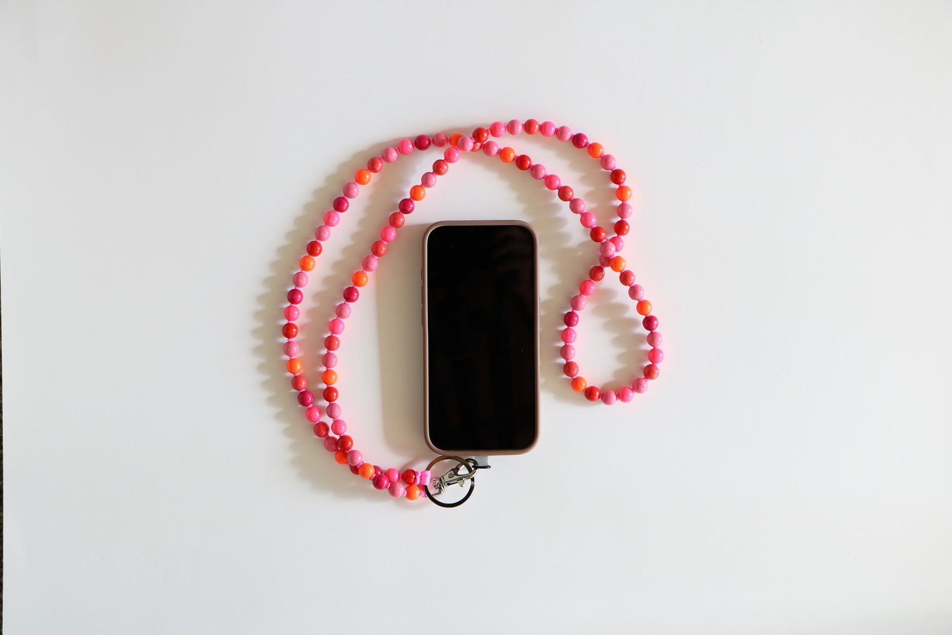 Phone with a multicolored pink wooden beaded lanyard attached. Made by Ina Seifart, the lanyard is on a white background with light pink, dark pink, red, and orange beads.