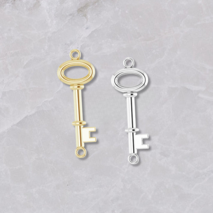 Two permanent jewelry connectors on a light marble background. Key-shaped, the left is 14k yellow gold and the right is sterling silver.