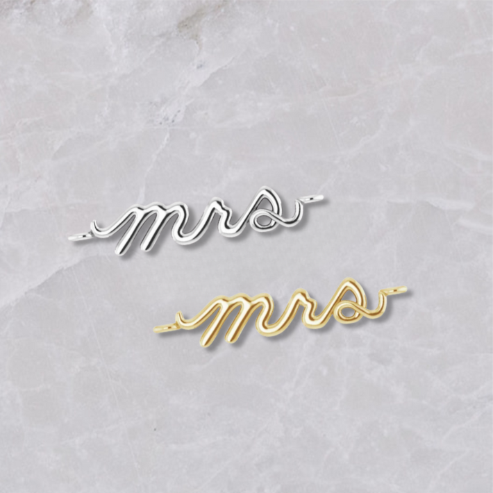 Permanent jewelry connectors with the word 'Mrs' shown in sterling silver and 14k yellow gold on a marble background. Perfect for newlyweds. 