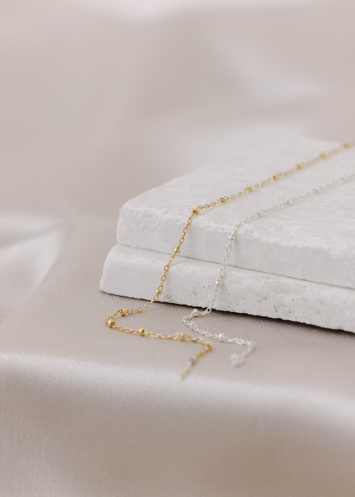 Two permanent jewelry links on a marble background and satin sheet. Dainty chain and beads in a pattern, with gold-filled on the left and sterling silver on the right. 