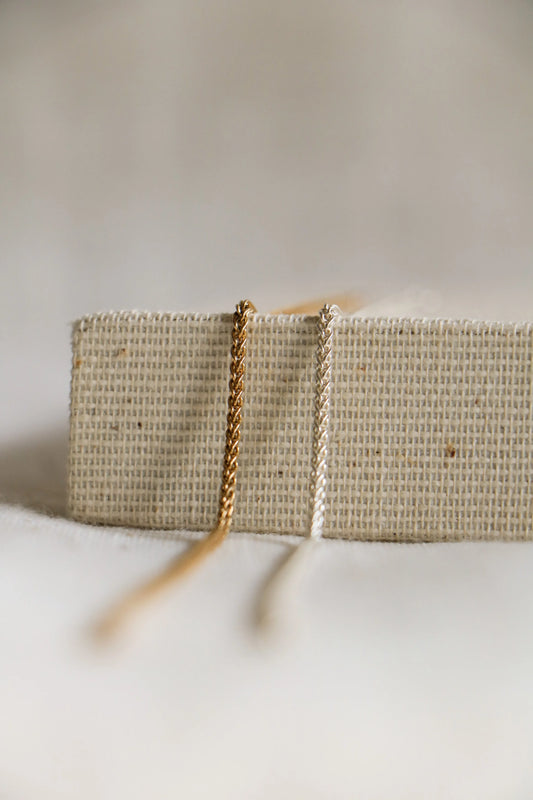 Two permanent jewelry chains close-up, laying on a woven holder. They are in a 'wheat' twisted shape, and shown in gold-filled on the left, and sterling silver on the right.