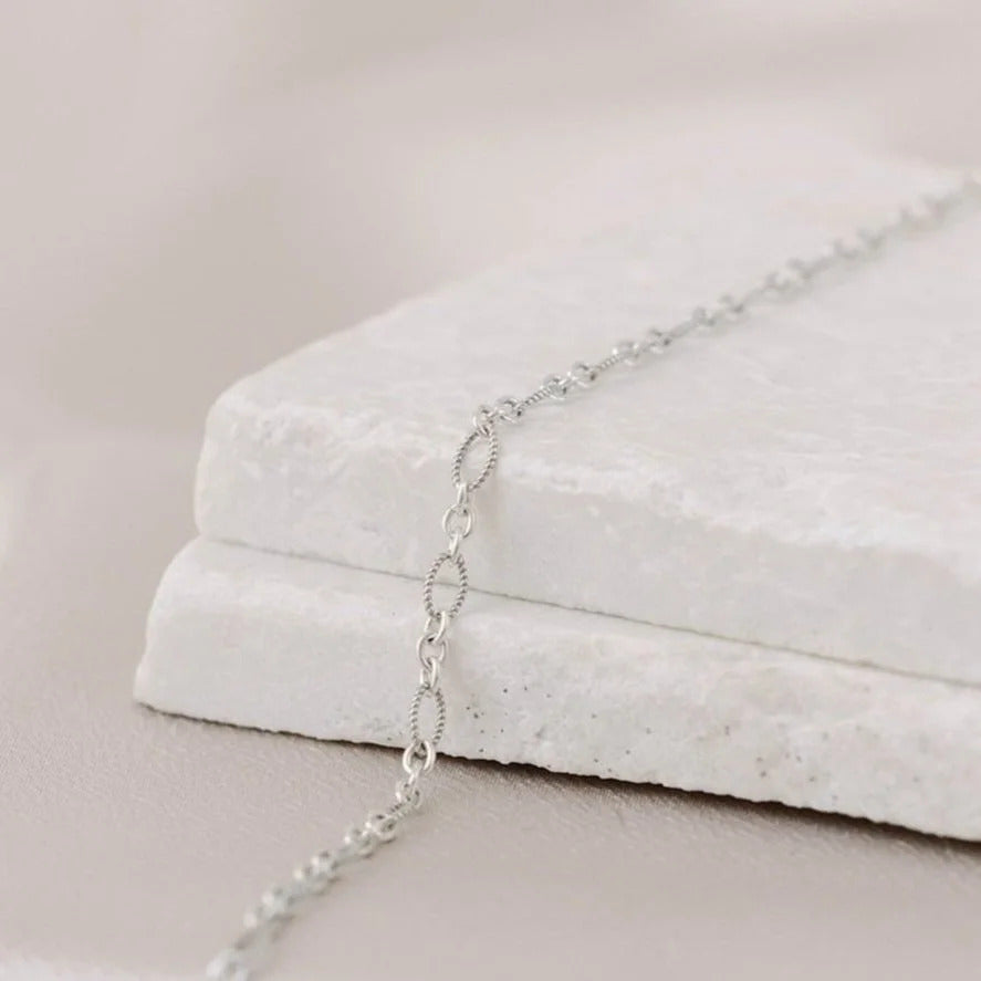 Statement permanent jewelry link called 'Twisted 3x1 Oval Link' shown in sterling silver on a white stone and satin background. The links are circular connectors, followed by twisted metal oval-shaped links. 