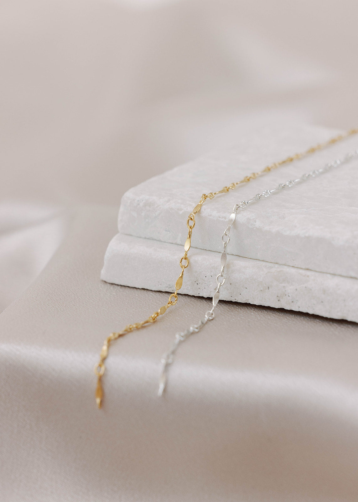Gold-filled and sterling silver permanent jewelry chains next to each other on a flat white stone with a satin background. The links have twisted filled bars followed by twisted oval links in a pattern.