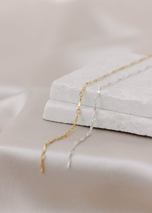 Gold-filled and sterling silver permanent jewelry chains next to each other on a flat white stone with a satin background. The links have twisted filled bars followed by twisted oval links in a pattern.
