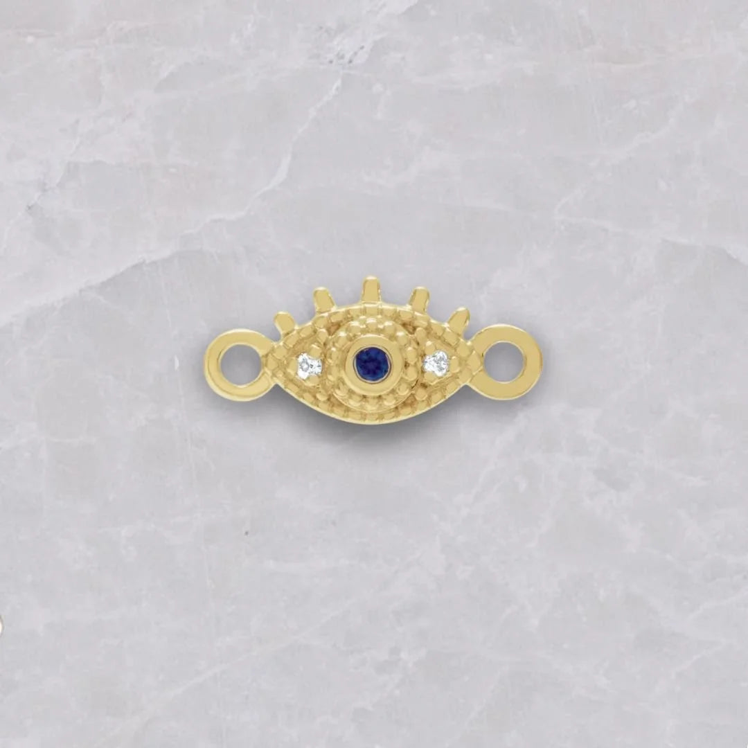 14k gold evil eye connector shown on a white marble background. The connector is an evil eye with loops on the side, 1 natural diamond on each side of the eye, and sapphire as the pupil. 